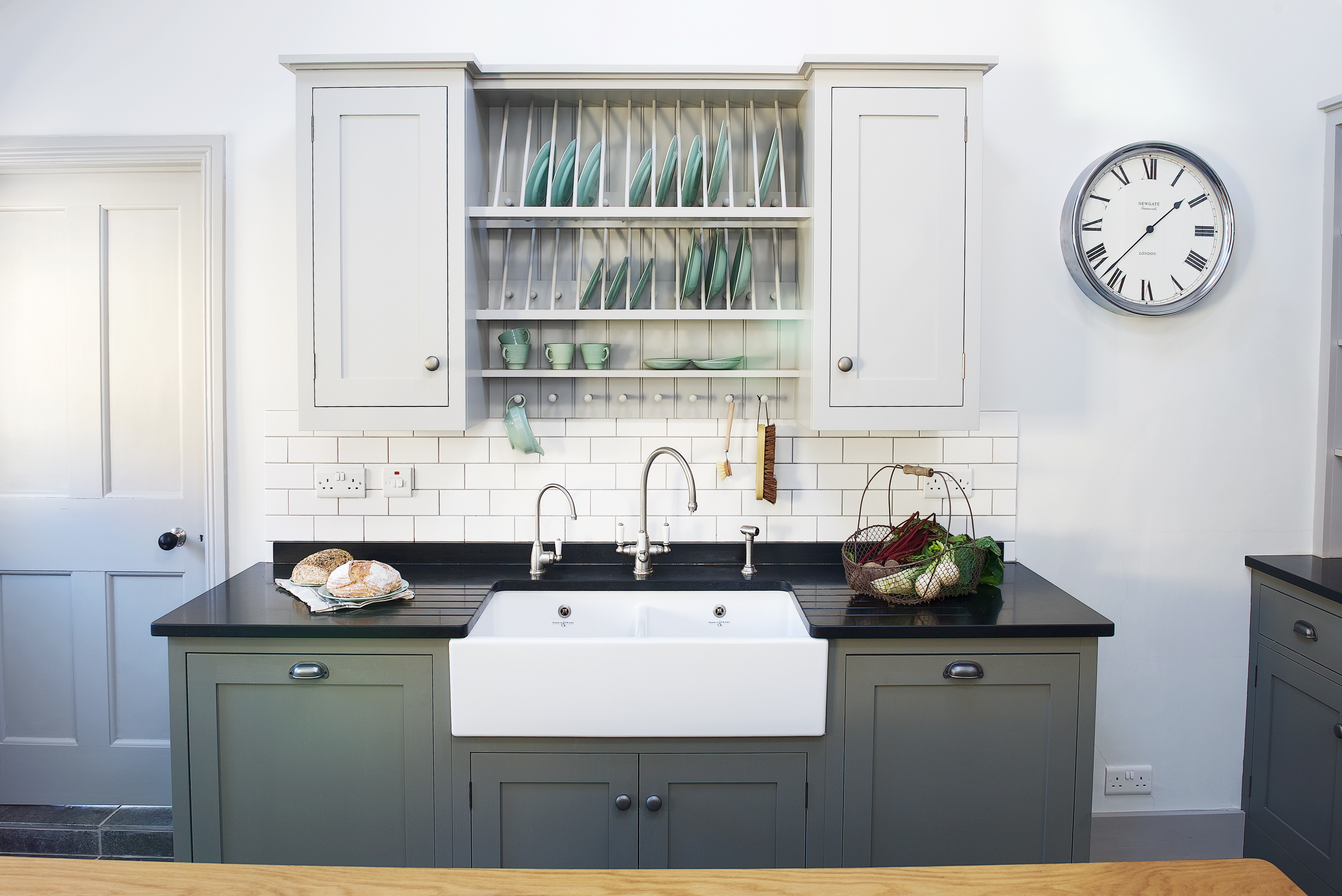 Grey and green shaker kitchen with pewter hardware and a double butler sink with black quartz worktops and a plate rack wall cabinet.