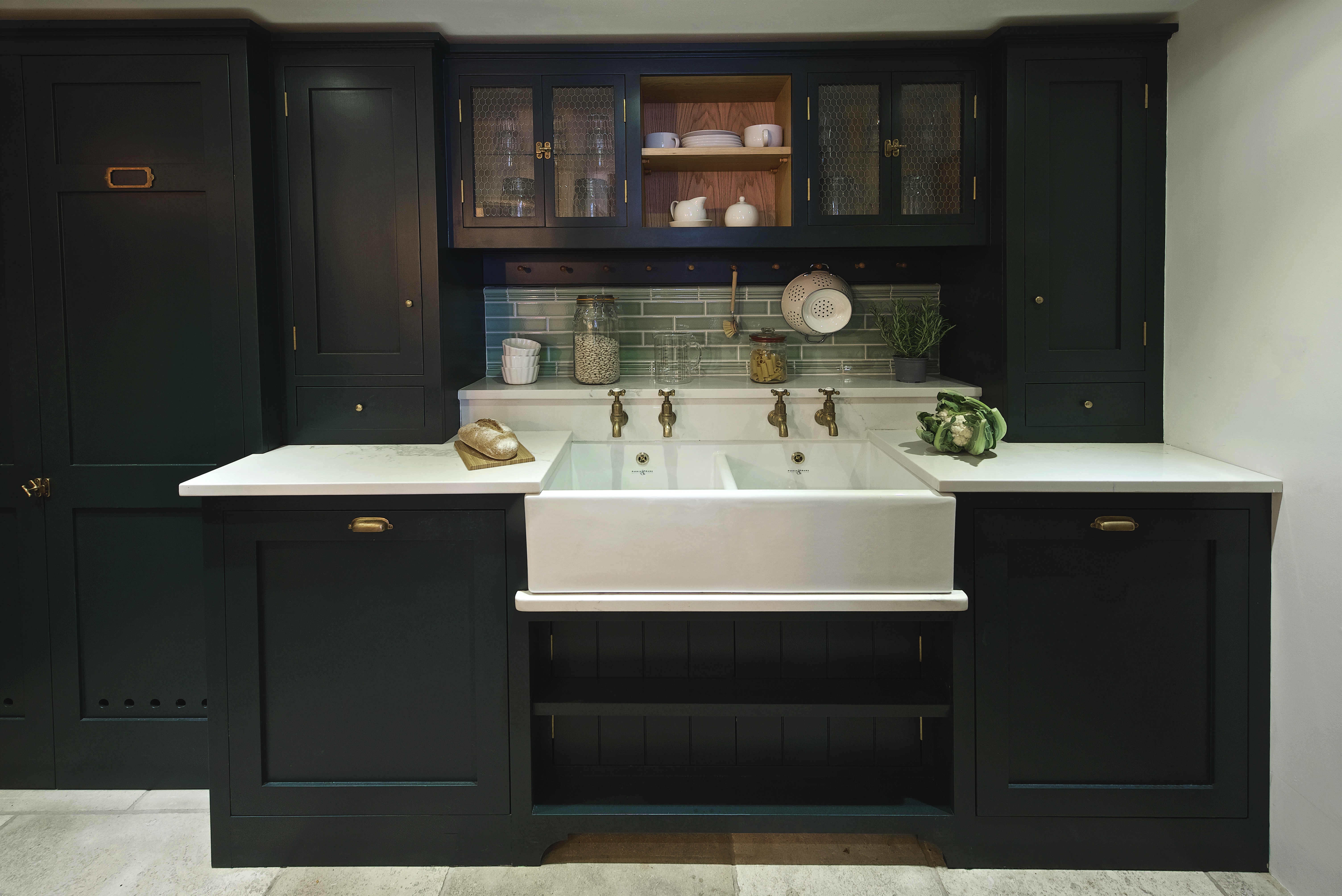 Dark blue shaker kitchen with white quartz worktops and a double belfast sink with brass hot and cold taps and green Fired Earth splashback tiles.