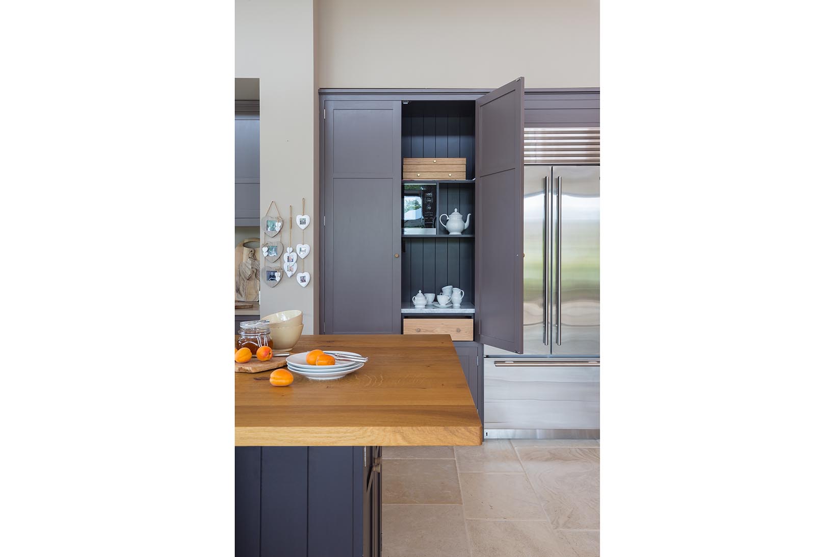 Island oak worktop close up with open tea and toast cabinet painted in Clay next to Subzero double fridge in stainless steel.