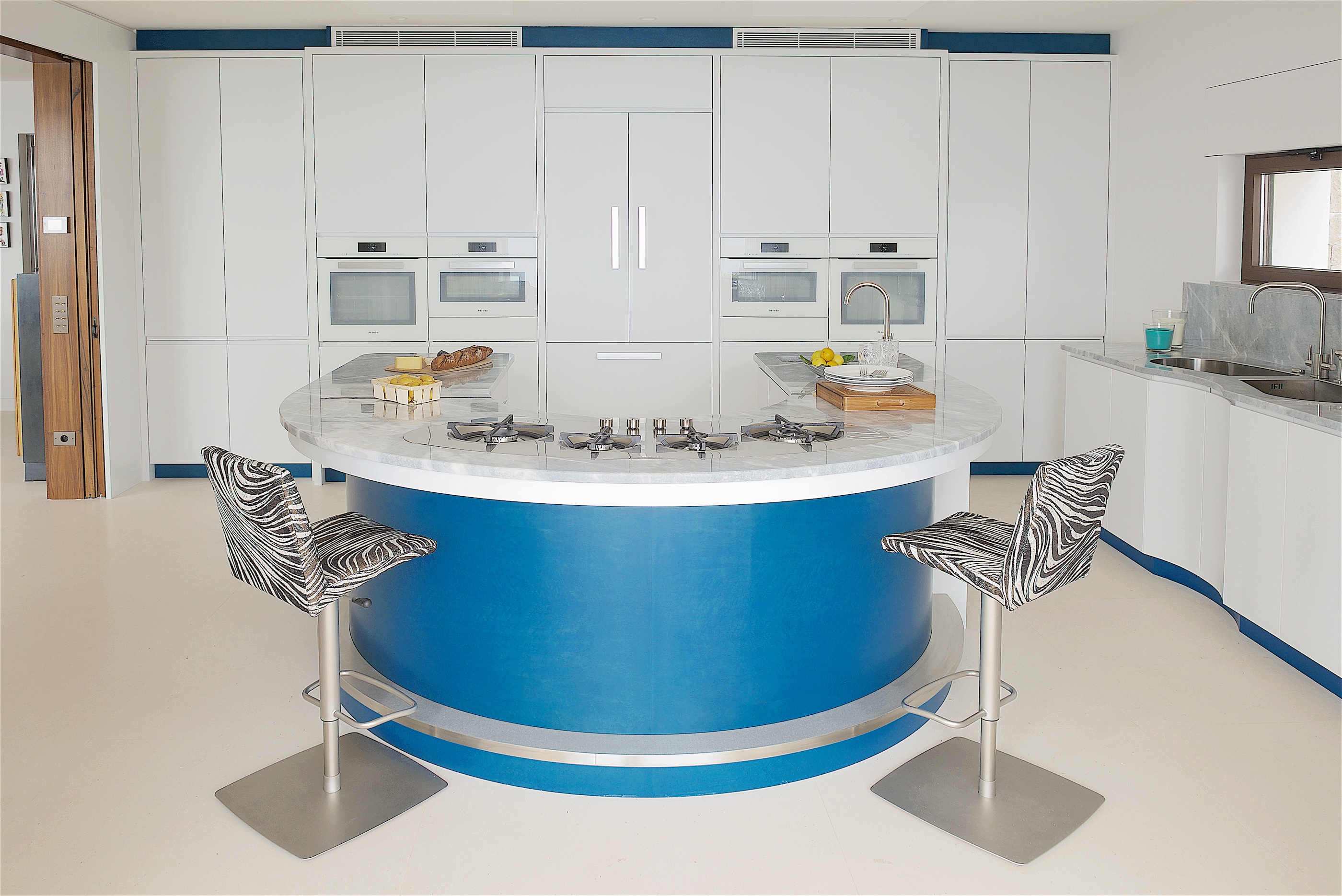 Contemporary blue curved kitchen island with Barazza Tao hob and zebra print kitchen stools with white kitchen cabinets and under cabinet lighting.
